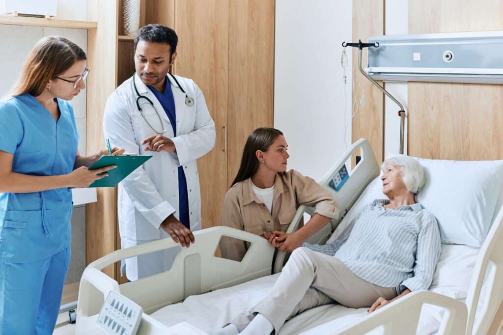 Doctor with nurse making round of hospital ward with sick elderly woman lying on bed. Bedside consultation